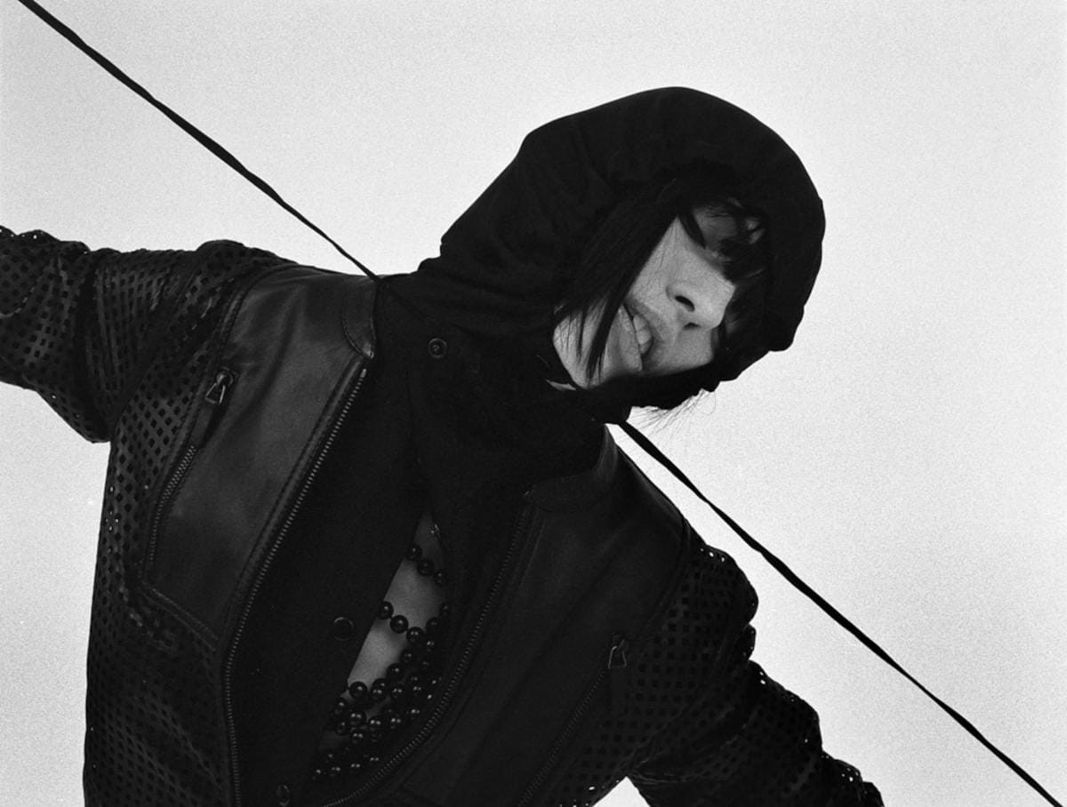 Woman wearing all black clothing with a hood, tilting her body to the right and pulling on the drawstrings