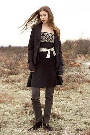A woman wearing a black dress with jacket and a white belt on a beach with dry grasses in winter