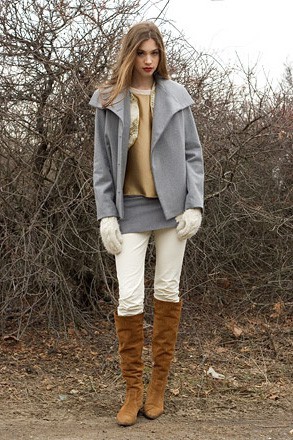 A woman wearing a gray jacket and skirt, with a light brown top and knee-high light brown boots in front of leafless small trees and bushes on a beach in winter