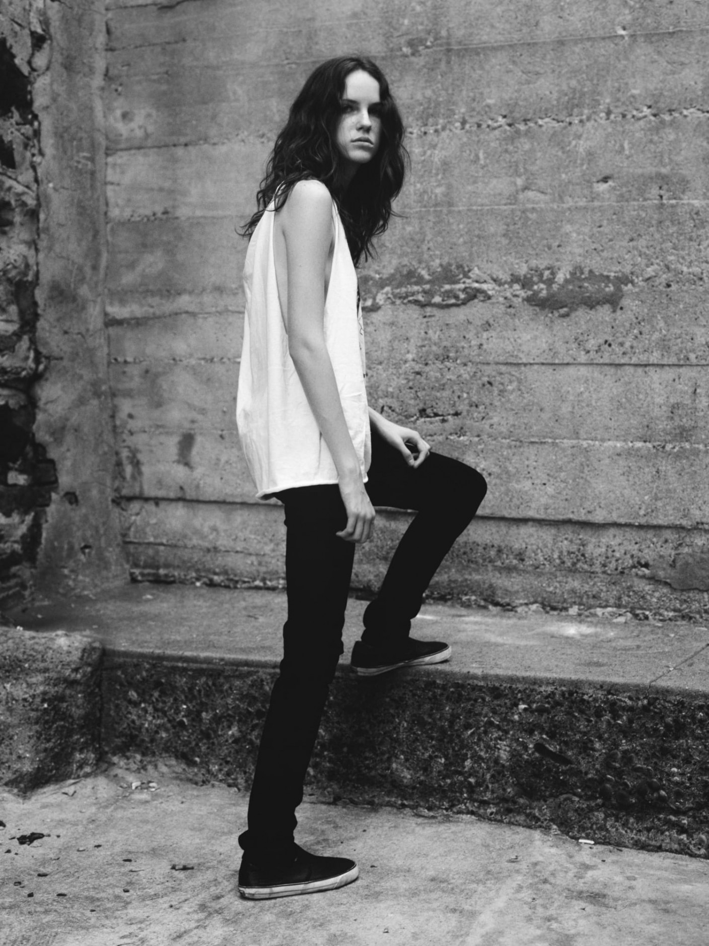 A model wearing a white cut off shirt and black jeans. One leg is raised up on a concrete ledge.