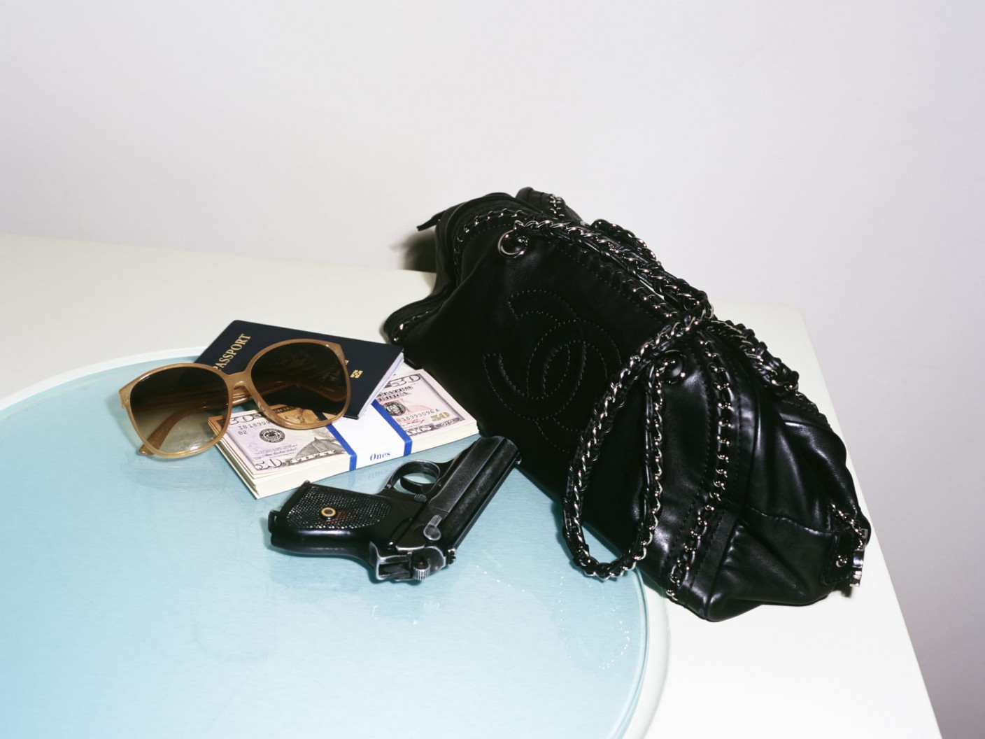 A handbag, a fake gun, a stack of fake bills, a passport, and sunglasses on a table