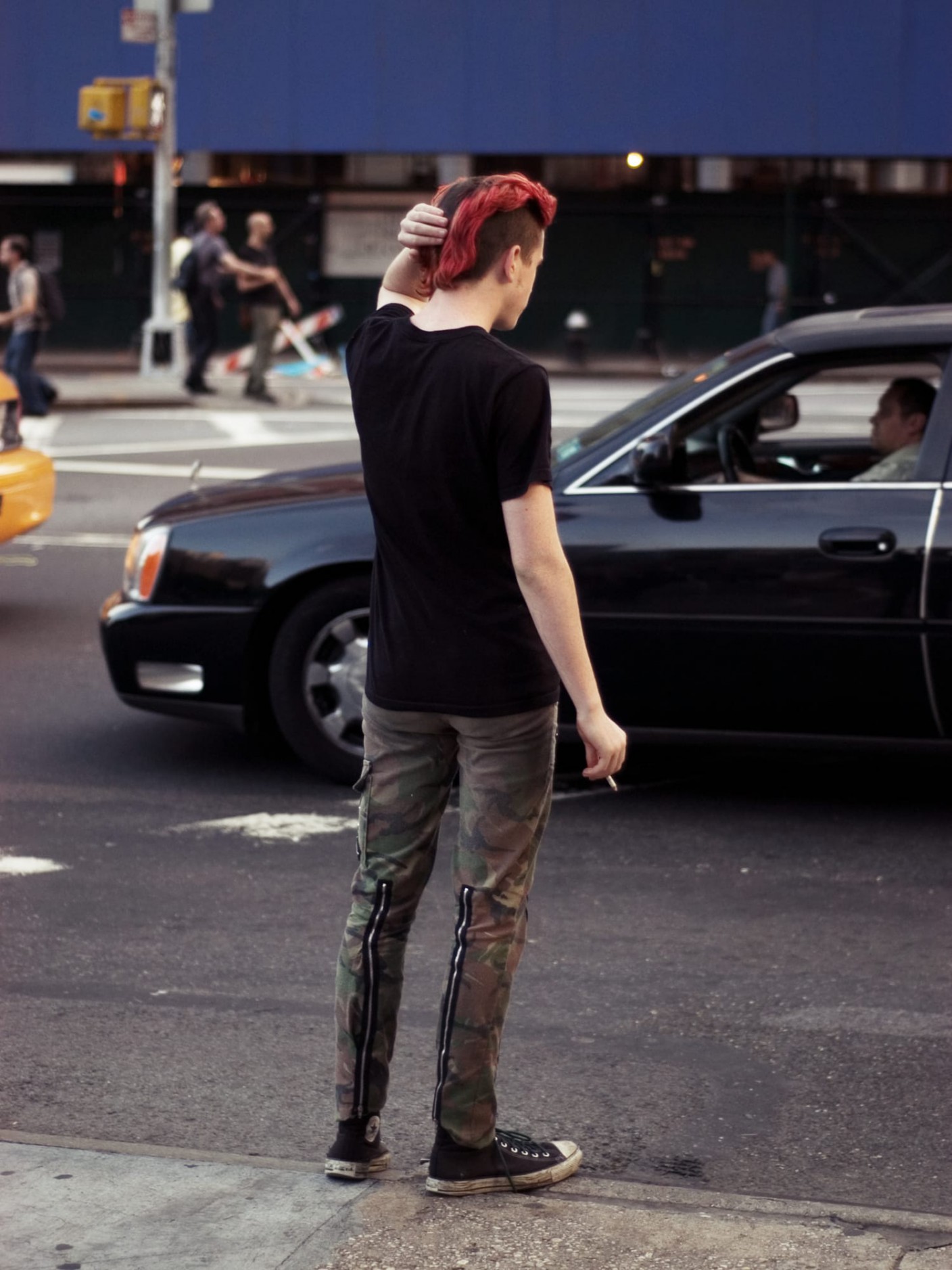 Portrait of a person on a sidewalk, with red dyed hair, a black shirt, and camouflage pants