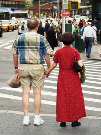 Portrait of a couple on a sidewalk, holding hands. One person is wearing a plaid shirt and the other is wearing a long red dress