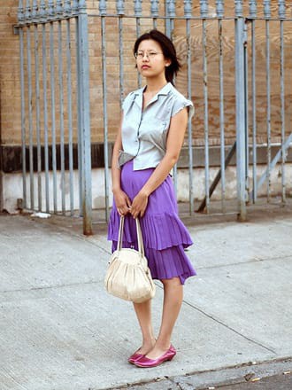 Portrait of a person in front of a light gray iron fence, wearing a purple skirt and carrying a beige purse