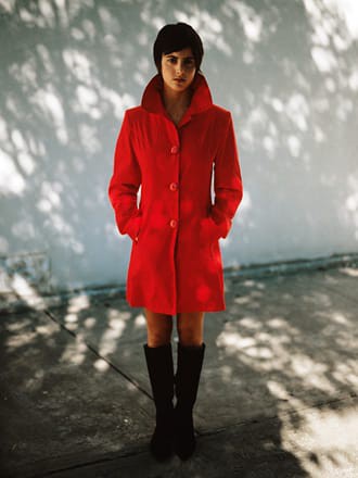 Portrait of a person wearing a red overcoat, under the shade of a tree, against a white wall